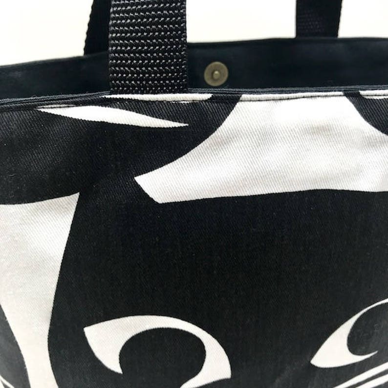Puede incluir: Bolsa de tela negra y blanca con un dise&ntilde;o de gato. La bolsa tiene asas negras y una cara de gato blanca con ojos grandes.