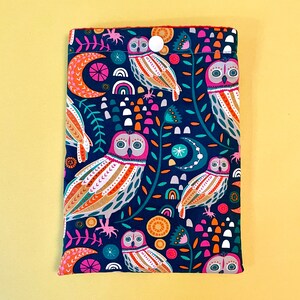 May include: A navy blue fabric book sleeve featuring a colorful owl pattern. The design includes owls, moons, rainbows, and floral elements in shades of pink, orange, teal, and white. A white button secures the top flap.
