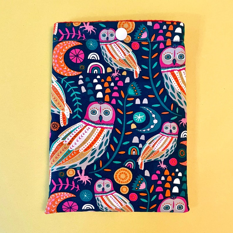 May include: A navy blue fabric book sleeve featuring a colorful owl pattern. The design includes owls, moons, rainbows, and floral elements in shades of pink, orange, teal, and white. A white button secures the top flap.