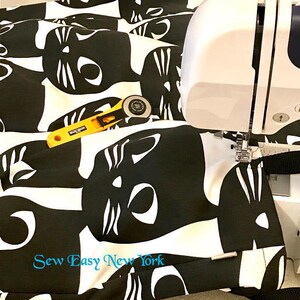 Puede incluir: Tela negra y blanca con un patr&oacute;n repetido de caras de gato. La tela se est&aacute; cosiendo en una m&aacute;quina de coser blanca. El texto "Sew Easy New York" est&aacute; impreso en la tela.