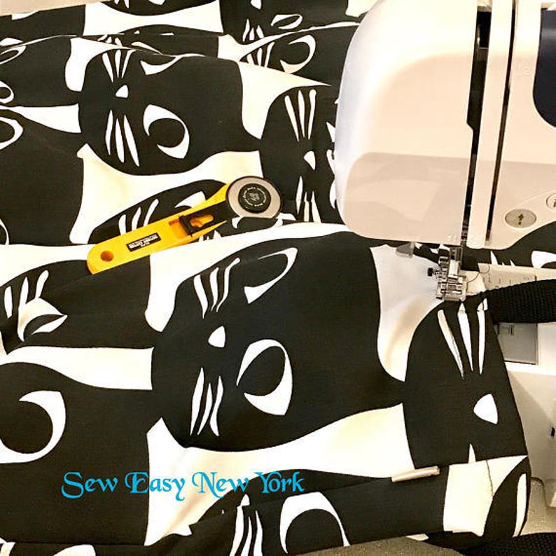 Puede incluir: Tela negra y blanca con un patr&oacute;n repetido de caras de gato. La tela se est&aacute; cosiendo en una m&aacute;quina de coser blanca. El texto "Sew Easy New York" est&aacute; impreso en la tela.