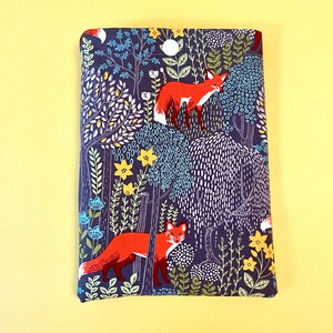 May include: A fabric book cover with a forest scene featuring red foxes, trees, and flowers. The background is a dark blue with white, yellow, and light blue accents. The cover is rectangular and appears to be made of cotton or a similar material.
