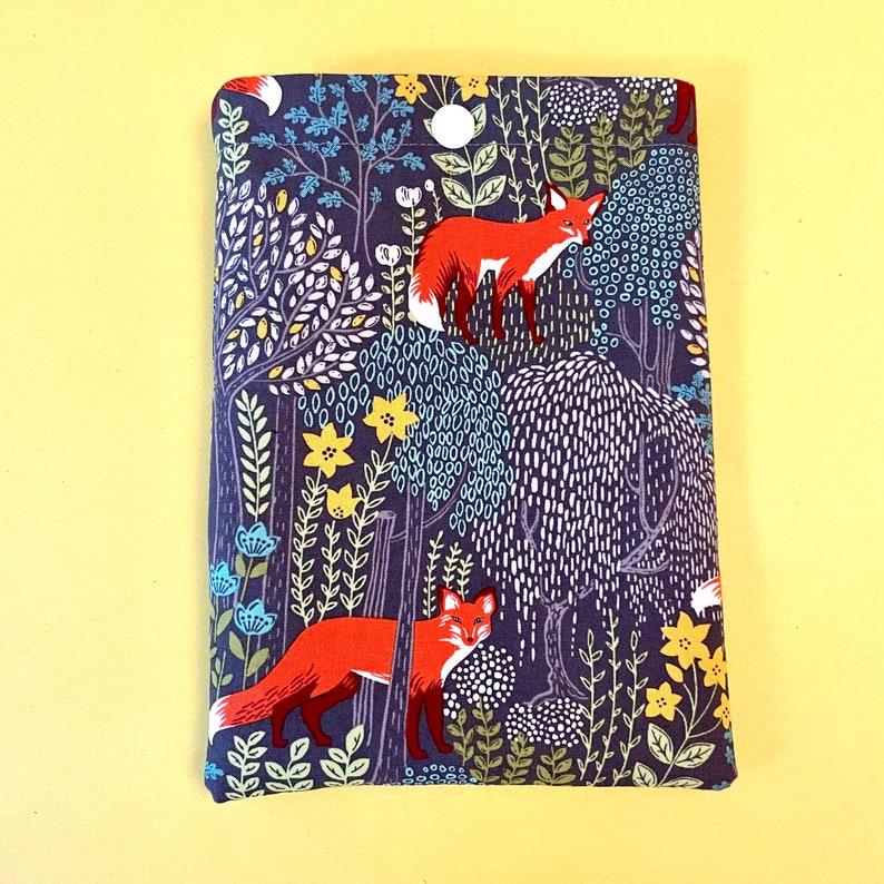 May include: A fabric book cover with a forest scene featuring red foxes, trees, and flowers. The background is a dark blue with white, yellow, and light blue accents. The cover is rectangular and appears to be made of cotton or a similar material.