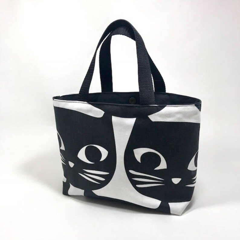 Puede incluir: Bolso de mano negro y blanco con un patr&oacute;n repetido de caras de gatos negros. El bolso tiene dos asas negras.