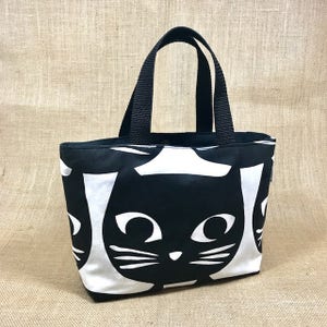 Puede incluir: Bolsa de tela negra y blanca con un patr&oacute;n repetido de caras de gatos negros. La bolsa tiene asas negras.