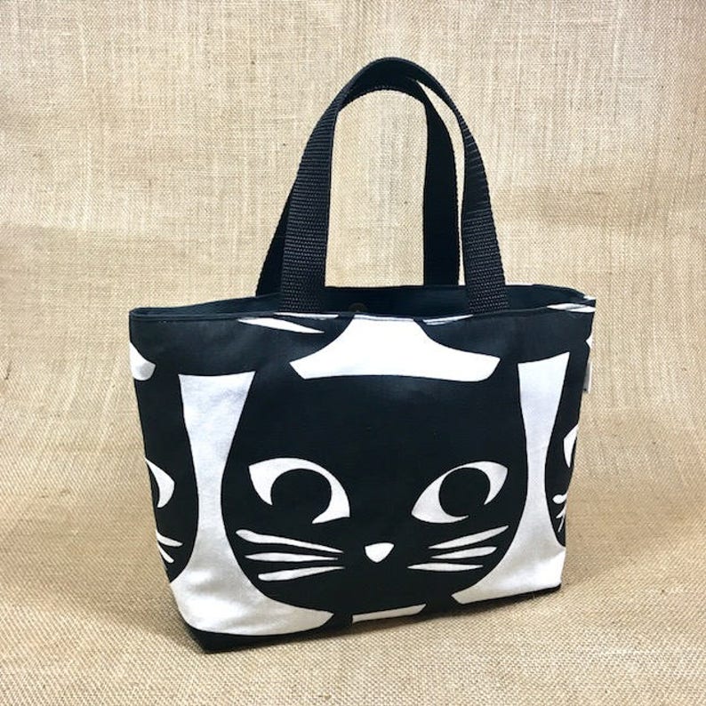 Puede incluir: Bolsa de tela negra y blanca con un patr&oacute;n repetido de caras de gatos negros. La bolsa tiene asas negras.