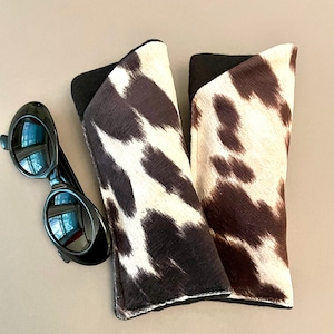 May include: Two brown and white cow print fabric eyeglass cases. The cases are lined with black fabric.