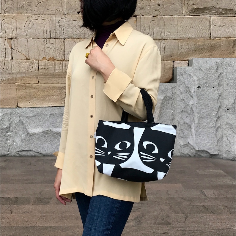Puede incluir: Una bolsa de mano de lona negra y blanca con un dise&ntilde;o de cara de gato. La bolsa tiene dos asas y la lleva una persona que lleva una camisa amarilla clara y pantalones vaqueros azules.