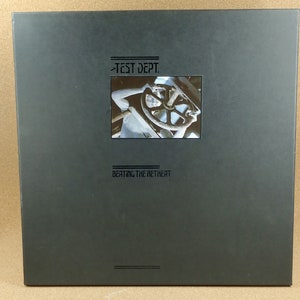 Può includere: Una copertina di un album di vinile nero con una foto di un ingranaggio metallico e il testo "Test Dept. Beating the Retreat" in bianco.