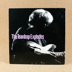 Può includere: Una foto in bianco e nero di una persona che suona la chitarra. Il testo "The Teardrop Explodes" è stampato in viola sulla parte anteriore del disco.