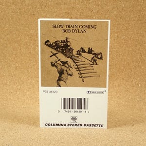 May include: A vintage cassette tape with a black and white illustration of a train on the front. The text "Slow Train Coming Bob Dylan" is printed on the front of the cassette tape. The cassette tape is labeled "Columbia Stereo Cassette" and has a barcode and the text "PCT 36120" printed on the back.
