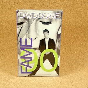 May include: A cassette tape case with a black and white photo of David Bowie on the front. The case is purple and green with the text "FAME" in large purple letters and "DAVID BOWIE" in smaller white letters.