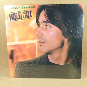 May include: A vinyl record album cover featuring a portrait of Jackson Browne. The album title "Hold Out" is printed in bold, white letters on a black background. The background is a blurry image of a sunset.
