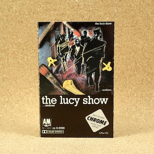 May include: A cassette tape case with a black background and white text that reads "the lucy show...undone". The front of the case features a black and white illustration of four people standing in a room with paintbrushes and other art supplies. The text "A&M on BASF CHROME MUSIC THAT SHINES" is also visible on the case.