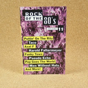 Può includere: Una custodia per CD intitolata "Rock of the 80's Volume 11" con uno sfondo astratto viola e bianco. La copertina elenca titoli di canzoni come "Funky Town" e "Men Without Hats".