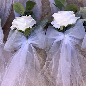 May include: White rose bouquets wrapped in white tulle with a white bow and a rhinestone embellishment. The bouquets are perfect for wedding decor or bridal party gifts.