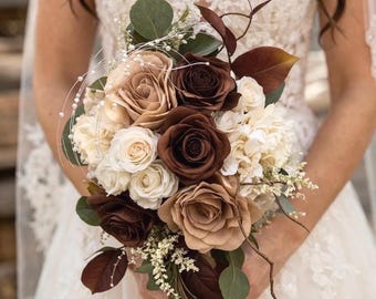 Puede incluir: Un ramo de novia con rosas en tonos marrón, crema y beige. El arreglo incluye vegetación y detalles decorativos, sostenido por una persona con un vestido de encaje blanco.