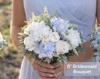 Könnte beinhalten: Ein 20 cm Brautjungfernstrauß mit weißen Rosen, blauen Hortensien und weißen Dahlien. Der Strauß ist mit grünem Laub akzentuiert. Der Strauß wird von einer Person in einem hellblauen Kleid gehalten. Eine schöne Hochzeitsanordnung.