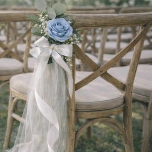 Può includere: Sedie in legno decorate con una rosa blu, verde e nastro e tulle bianchi. Le sedie sono allestite per un evento all'aperto, forse un matrimonio. Le sedie hanno una finitura in legno chiaro e sedili imbottiti.