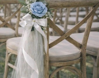 Könnte beinhalten: Holzstühle, verziert mit einer blauen Rose, Grünzeug und weißem Band und Tüll. Die Stühle sind für eine Outdoor-Veranstaltung, möglicherweise eine Hochzeit, aufgestellt. Die Stühle haben eine helle Holzoberfläche und gepolsterte Sitze.