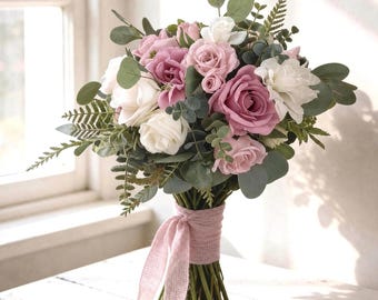 Puede incluir: Un ramo de flores con rosas malvas y blancas, intercaladas con hojas de eucalipto verdes y frondas de helecho. Los tallos están reunidos y atados con una cinta de tela rosa claro texturizada que cuelga en dos extremos largos.