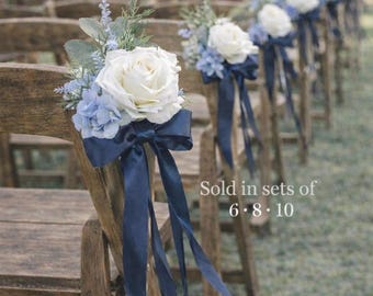 Könnte beinhalten: Holzstühle, verziert mit weißen Rosen, blauen Hortensien und marineblauen Bändern. Die Blumenarrangements sind an den Stuhlrücken befestigt und bilden eine dekorative Präsentation für eine Hochzeit oder eine besondere Veranstaltung.