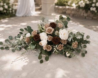 Puede incluir: Un arreglo floral que presenta una mezcla de rosas blancas, beige y marrón oscuro intercaladas con hojas de eucalipto verdes y pequeñas flores blancas. Las flores están dispuestas en un grupo horizontal alargado con follaje extendido.