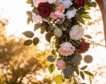 Puede incluir: Un arreglo floral con rosas de color rojo intenso, rosa suave y blanco, mezcladas con pequeñas flores blancas de relleno y hojas verdes de eucalipto. Las flores están agrupadas y cuelgan de una estructura de arco de madera curvada.