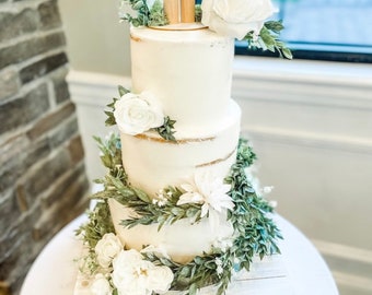 Op de afbeelding: Een drielaagse witte bruidstaart versierd met groen en witte bloemen. De bovenste laag heeft een houten topper met twee kleine houten figuurtjes.