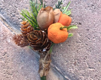 Puede incluir: Un prendedor decorativo con dos pequeñas calabazas naranjas, un tulipán de tela marrón, dos piñas marrones y follaje verde. Los tallos están envueltos en cuerda natural. El texto PUMPKIN PATCH está impreso en negro en la parte inferior.