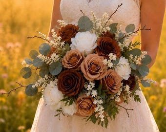 May include: A floral bouquet featuring a mix of white, tan, and dark brown roses, accented with green eucalyptus leaves, small white filler flowers, and thin brown twigs. The arrangement is held in front of a white lace dress with a cap sleeve design.