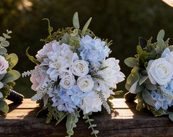 Könnte beinhalten: Drei Blumensträuße mit weißen, hellblauen und rosa Blüten, akzentuiert mit grünem Laub, liegen auf einer Holzoberfläche. Die Sträuße sind in einem natürlichen, organischen Stil angeordnet, ideal für Hochzeiten oder besondere Anlässe.