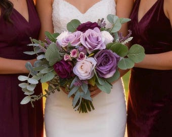 Könnte beinhalten: Ein Brautstrauß mit lila und lavendelfarbenen Rosen, akzentuiert mit Zweigen von Grün. Der Strauß wird von einer Person in einem weißen Hochzeitskleid gehalten. Zwei Personen in bordeauxfarbenen Kleidern stehen auf beiden Seiten.