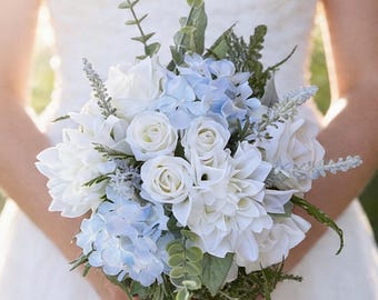 Könnte beinhalten: Ein Brautstrauß mit weißen Rosen, Dahlien und blauen Hortensien. Das Arrangement umfasst grünes Laub und wird von einer Person in einem weißen Kleid gehalten. Die Blumen sind eng angeordnet und bilden eine volle, runde Form.
