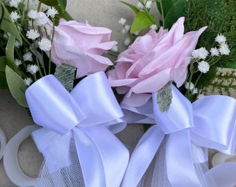 Könnte beinhalten: Nahaufnahme von zwei Blumengestecken mit hellrosa Rosen, weißen Schleifen und zartem weißem Netz. Grüne Blätter und kleine weiße Blüten akzentuieren die Rosen. Die Schleifen sind aus weißem Satinband.