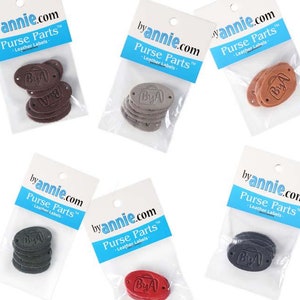 May include: Five packages of leather labels for purses, each package contains six oval-shaped labels with the text "ByA" and a small suitcase icon. The labels are in different colors: brown, gray, black, red, and orange.