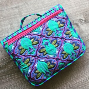 May include: A quilted zippered pouch with a teal, purple and green floral pattern. The pouch has a pink zipper and a teal handle.