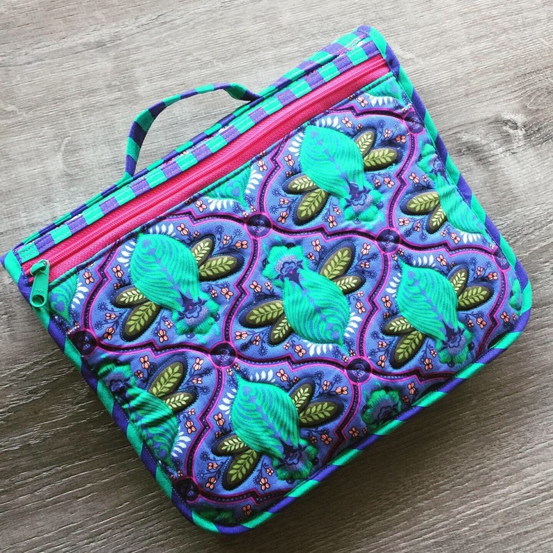 May include: A quilted zippered pouch with a teal, purple and green floral pattern. The pouch has a pink zipper and a teal handle.