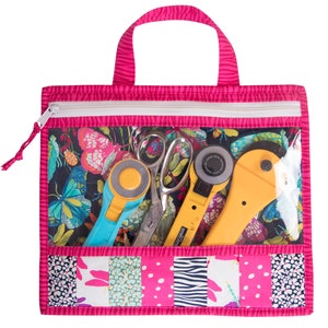 Puede incluir: Una bolsa de mano rosa con cremallera y un bolsillo frontal transparente de vinilo. El bolsillo contiene un cortador rotatorio, tijeras y una regla azul. La bolsa est&aacute; hecha de tela de patchwork con una variedad de patrones, incluyendo mariposas, lunares y rayas.