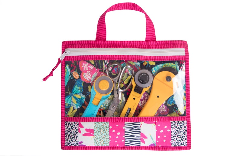 Puede incluir: Una bolsa de mano rosa con cremallera y un bolsillo frontal transparente de vinilo. El bolsillo contiene un cortador rotatorio, tijeras y una regla azul. La bolsa est&aacute; hecha de tela de patchwork con una variedad de patrones, incluyendo mariposas, lunares y rayas.