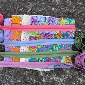 May include: Assortment of colorful zipper tape in pink, purple, green, and blue. The tape is arranged in a stack with fabric scraps underneath.