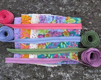 Puede incluir: Surtido de cintas de cremallera de colores en rosa, morado, verde y azul. La cinta está dispuesta en una pila con recortes de tela debajo.