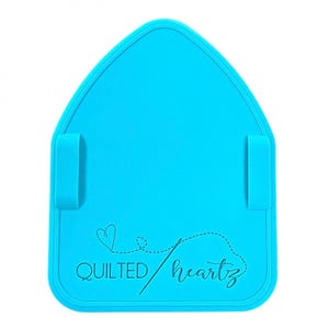 May include: A bright turquoise sewing mat with a unique house shape. The mat features two attached loops and the words "QUILTED heartz" in a cursive font with a heart design. The mat is designed for quilting and sewing projects.