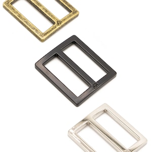 May include: Three metal rectangle sliders in antique brass, black, and silver. Each slider has a rectangular frame with a central bar, and a small logo is visible on each. The sliders are displayed on a white background.