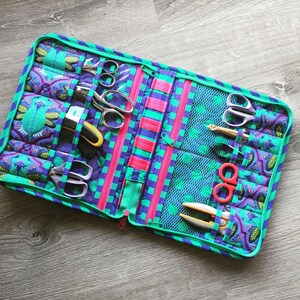 May include: A teal and purple zippered pouch with multiple pockets and compartments. The pouch is filled with various sewing tools, including scissors, needles, and thread.