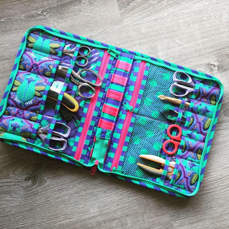 May include: A teal and purple zippered pouch with multiple pockets and compartments. The pouch is filled with various sewing tools, including scissors, needles, and thread.