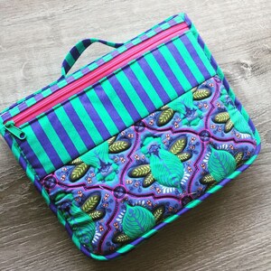 May include: A teal and purple striped fabric pouch with a pink zipper and a green and purple floral patterned fabric lining. The pouch has a handle on top.
