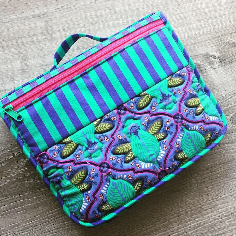 May include: A teal and purple striped fabric pouch with a pink zipper and a green and purple floral patterned fabric lining. The pouch has a handle on top.