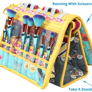 May include: Yellow and blue makeup brush holder with multiple compartments and a zipped closure. The holder has a mesh pocket and a strap. The text "Running With Scissors PBA272" and "Take A Stand PBA273" are visible on the holder.