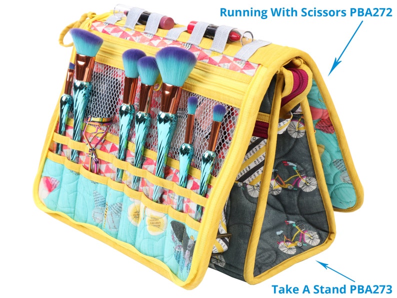 May include: Yellow and blue makeup brush holder with multiple compartments and a zipped closure. The holder has a mesh pocket and a strap. The text "Running With Scissors PBA272" and "Take A Stand PBA273" are visible on the holder.